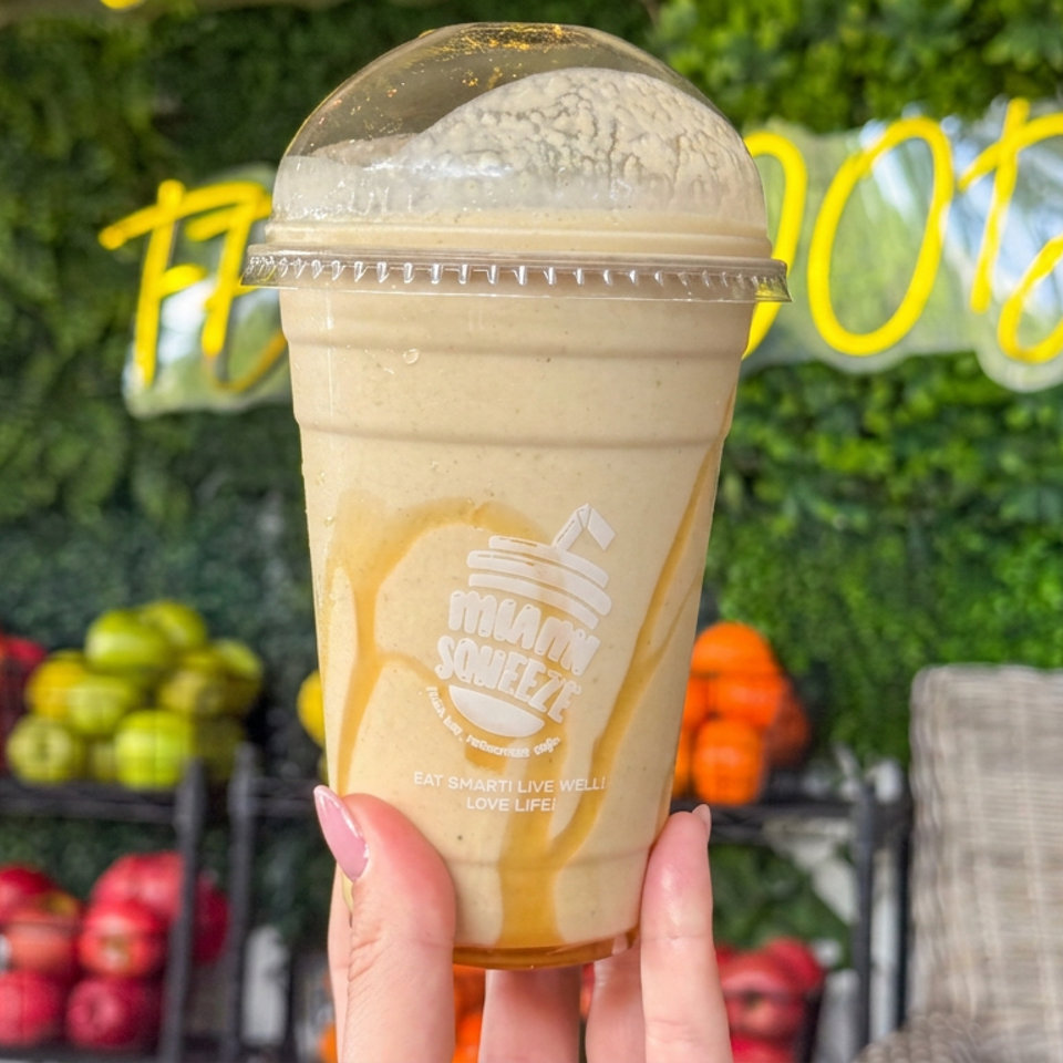 Hand holding a creamy blended drink in a branded cup, with colorful fresh produce displayed against a leafy green wall.