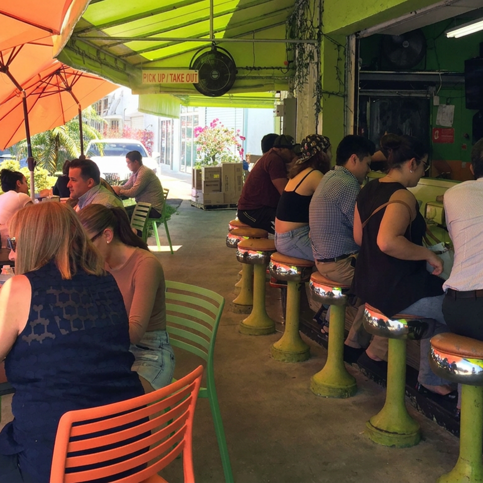 Busy colorful outdoor restaurant with orange umbrellas, green counter seating, and diners enjoying meals.